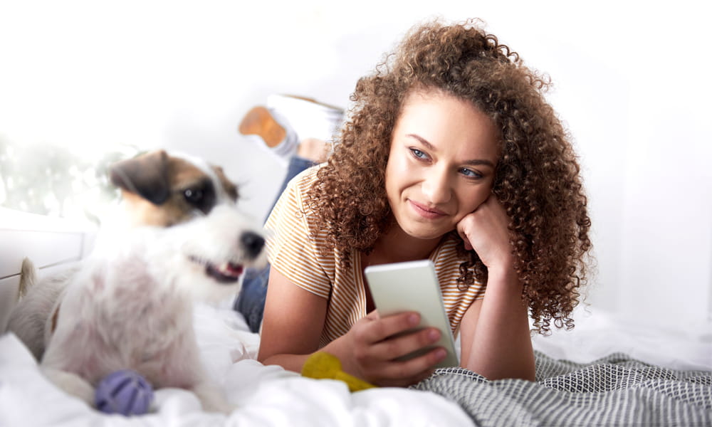 Woman holding her phone while on the bed with her dog